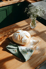 Fresh homemade rustic bread on a light wooden table, linen napkin, scattered flour, a vase of wildflowers, cozy quiet morning, slow living, homemade food, bakery, and rustic lifestyle concepts