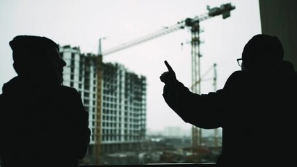 Silhouetted engineers discussing construction project with tower crane and building site in background showing teamwork planning, architecture development, urban infrastructure progress