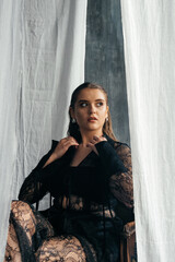 Confident woman in sheer black lace stands poised between white curtains, embodying strength, elegance, and modern gothic fashion
