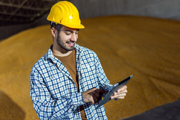 Young man stands in warehouse in front of large soybean pile holding tablet
