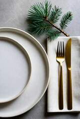 A minimalist holiday table setting with white plates, a crisp napkin, gold fork and knife, and a pine sprig centerpiece, conveying festive warmth and refined dining style.