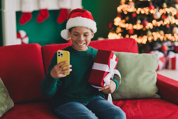 A cheerful boy holding a festive gift and enjoying holiday greetings on his smartphone in a cozy Christmas-themed room.