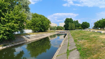 Fototapeta premium The river flows through the city in banks lined with concrete slabs. Nearby there are grassy lawns with trees reflecting in the water, a concrete fence and city buildings. Sunny summer weather