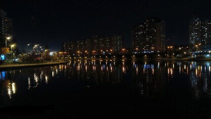 Night cityscape with lights reflecting on lake. Tall residential buildings illuminated at night with colorful streetlights reflecting on a calm urban lake in a peaceful setting