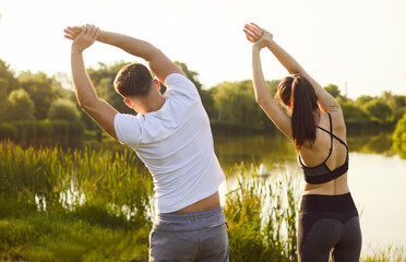 Sport exercise and healthy workout of active couple stretching body in side bends. Back view of two fit people in sportswear training at summer forest lake in morning. Weight loss, training concept