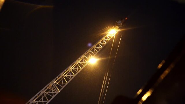 Crane silhouette pierces dark sky illuminated by sharp floodlights. Night atmosphere turns routine port work into striking visual drama