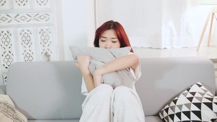 Young worried asian woman sitting on a sofa holding a pillow, displaying sadness and contemplation, likely reflecting on emotional struggles and mental health concerns