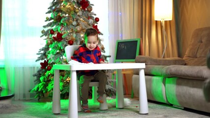 Lovely cheerful boy pulls the desk to the chair and sits down. Decorated Christmas tree at backdrop.