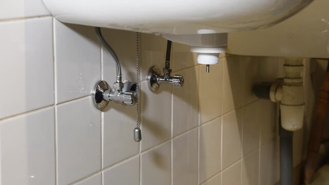 Experienced plumber performing routine maintenance under a white ceramic sink, unscrewing the drain plug to clear debris and resolve a bathroom blockage, demonstrating professional repair work