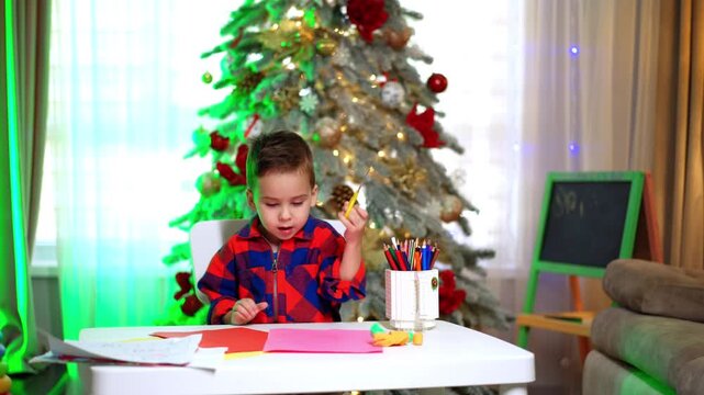 Cute 4-year-old toddler in checkered shirt doing art sitting at desk. Baby boy cuts color paper and chooses a pencil from a mug. Christmas tree at backdrop.