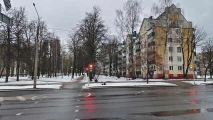 After a snowfall, the ground is covered with snow. Municipal services have cleared the roadway and sidewalks. Nearby there are traffic lights, buildings, lampposts and trees. Cloudy winter weather © Balser