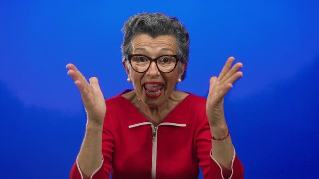Elderly woman with grey hair wearing glasses and a red top expressing surprise against a vibrant blue background.