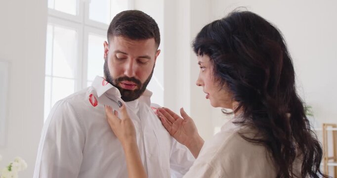 Couple fight over lipstick stain on husbands shirt. Angry woman asking questions of cheating man with red marks on clothes, quarrel over suspicions of affair and infidelity, betrayal and trust issues