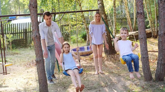 Parents and children having fun outdoors in backyard. Mom and Dad pushing boy and girl on swing at playground. Family vacation outside city, playground