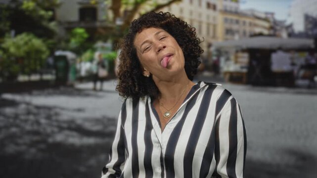 Hispanic middle aged woman in bright striped shirt sticks tongue out on sunlit urban street; playful fun.