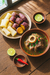Rustic overhead shot of traditional Indonesian beef bone marrow soup with cassava and sweet potatoes by a window on wooden table, fresh lime and chili garnish, generative AI