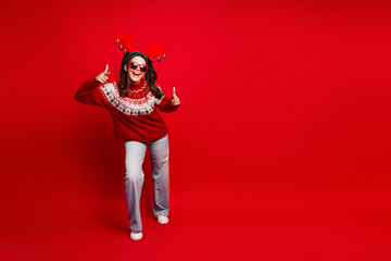Happy teen girl in red christmas sweater with reindeer headband posing against bold red background celebrating festive season and holiday joy with thumbs up and playful fashion