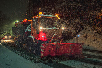 Snow plow truck working at night during snowfall. Illuminated winter street scene with vehicles and snowflakes.