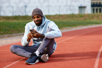 Smiling male athlete sits on a running track and checks his mobile phone during a relaxed break, wearing earbuds and sportswear.