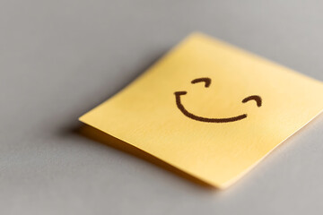 A cheerful yellow sticky note, featuring a simple hand-drawn smiling face in black marker, set against a neutral background, conveys positivity and a reminder to smile.