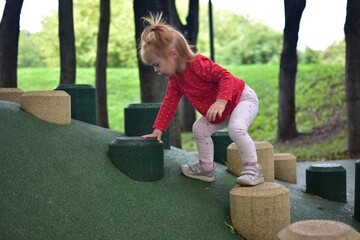 Toddler girl climbing steps on playground hill shaped structure outdoors, focused expression, wearing red shirt and pastel leggings, developing coordination and balance in natural park environment.