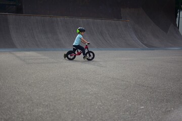 Young boy riding balance bike alone in skate park area with large ramp behind, active motion and learning balance outdoors in recreational sports zone.