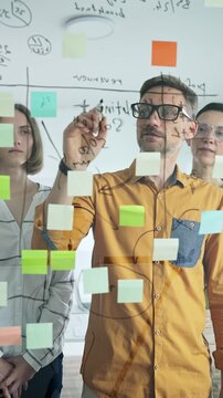 Professional business team brainstorming, sharing strategy on glass whiteboard with colorful sticky notes, working collaboratively in bright office space. Designers at work