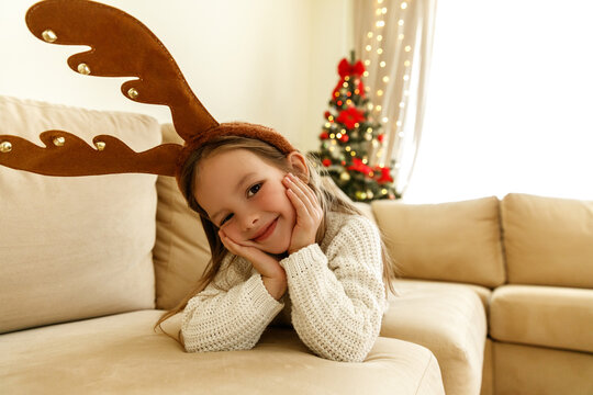Cute little girl in reindeer antlers smiling on beige sofa, resting her face on hands. Christmas tree with lights and red decorations glowing in background, festive holiday atmosphere.