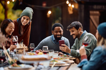 Happy men using cell phone during lunch party with friends on patio in autumn.