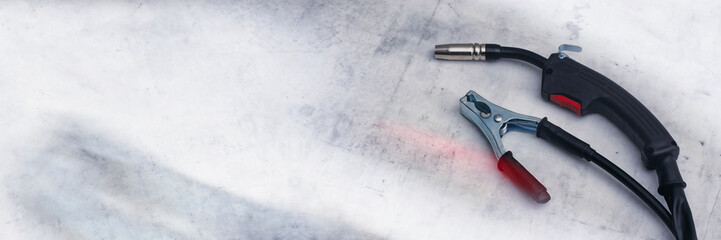 Close-up of professional MIG MAG welding torch welding gun and a copper ground clamp earth clamp resting on a metallic, scratched workbench or surface.