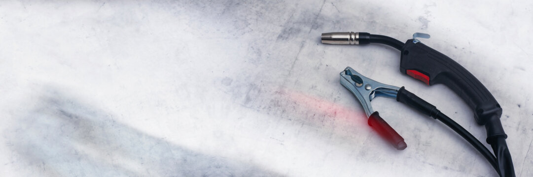 Close-up of professional MIG MAG welding torch welding gun and a copper ground clamp earth clamp resting on a metallic, scratched workbench or surface.