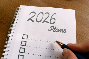 2026 Plans,  A spiral notebook on wooden desk with checkboxes for goals, planning, and future ideas for upcoming New Year., with grid pages and hand holding blue pen