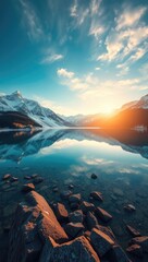 Serene mountain lake reflecting snowcapped peaks and a bright sunset with rocks in the foreground