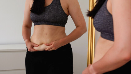 Woman checking her stomach in front of a mirror, holding belly skin fold. Concept of body image, self-acceptance, fitness progress, postpartum changes, and realistic health goals