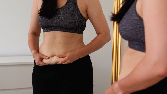 Woman checking her stomach in front of a mirror, holding belly skin fold. Concept of body image, self-acceptance, fitness progress, postpartum changes, and realistic health goals