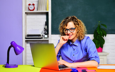 Fototapeta premium Smiling female teacher or businesswoman in glasses working with laptop at workplace. Student girl preparing for exam using laptop in classroom. Online courses, webinar, e-learning or virtual training.