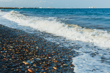 Waves gently lap against a rocky shore on a sunny day near a coastal city, with distant buildings glimmering in the sunlight and the horizon meeting the vibrant blue sky