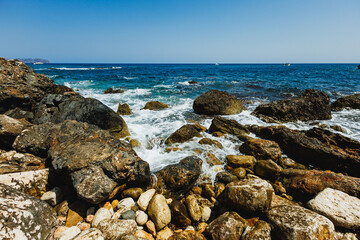 Ocean waves crash against rugged stones under a clear blue sky on a sunny day by the coast