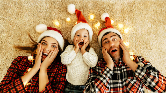 Amazed family lying on the carpet with glowing lights, parents and daughter in Santa hats holding their faces with joy, celebrating love, Christmas eve, and New Year.