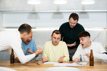 Fototapeta premium Upset worried man sitting at table in home kitchen with papers in hands, reading unexpected medical results with group of male friends solacing and comforting him ....