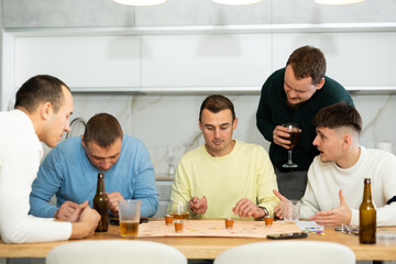 Cheerful young adults enjoying beer and wine immersed in board game with twist, where shot glasses of alcoholic drinks replacing traditional game pieces