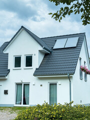 Modern white house with solar panels surrounded by greenery on a partly cloudy day near a community