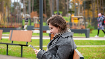 Woman Sitting on a Park Bench Using Her Smartphone in a Lively Green Space During Autumn