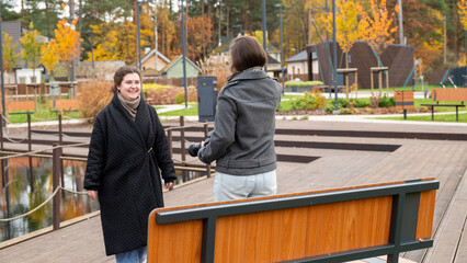 Two Friends Enjoy a Sunny Autumn Day at the Park Sharing Laughter and Conversation Near a Lake Surrounded by Colorful Trees