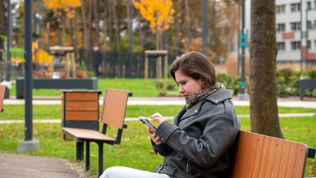 Young Person Sitting on a Bench in a Park During Autumn, Focused on Phone Screen While Surrounded by Colorful Trees - Powered by Adobe