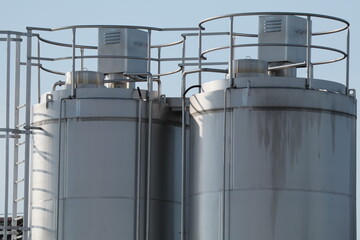 Two large industrial metal silos stand under a clear blue sky, their silver surfaces gleaming in the sunlight. The clean lines, pipes, and ladders highlight modern engineering and precision.