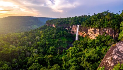 Naklejka premium Lush green forest valley with a waterfall cascading down a rock face beneath a bright, partly cloudy sky