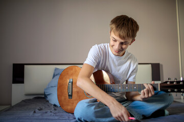 Creative teenage boy sitting on his bed playing an acoustic guitar and writing down lyrics or notes on a notepad