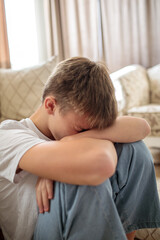 Sad and depressed teenage boy sitting on the floor with his head buried in his arms, expressing...