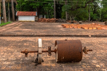 Old rusty soil compactor abandoned on a brick yard, surrounded by clay roof tiles and trees, showing obsolete rural technology replaced by modern agriculture.